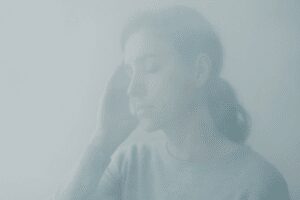 A soft-focus portrait of a woman gazing into the distance with a gentle hand to her temple, surrounded by a subtle mist, symbolizing migraine brain fog.